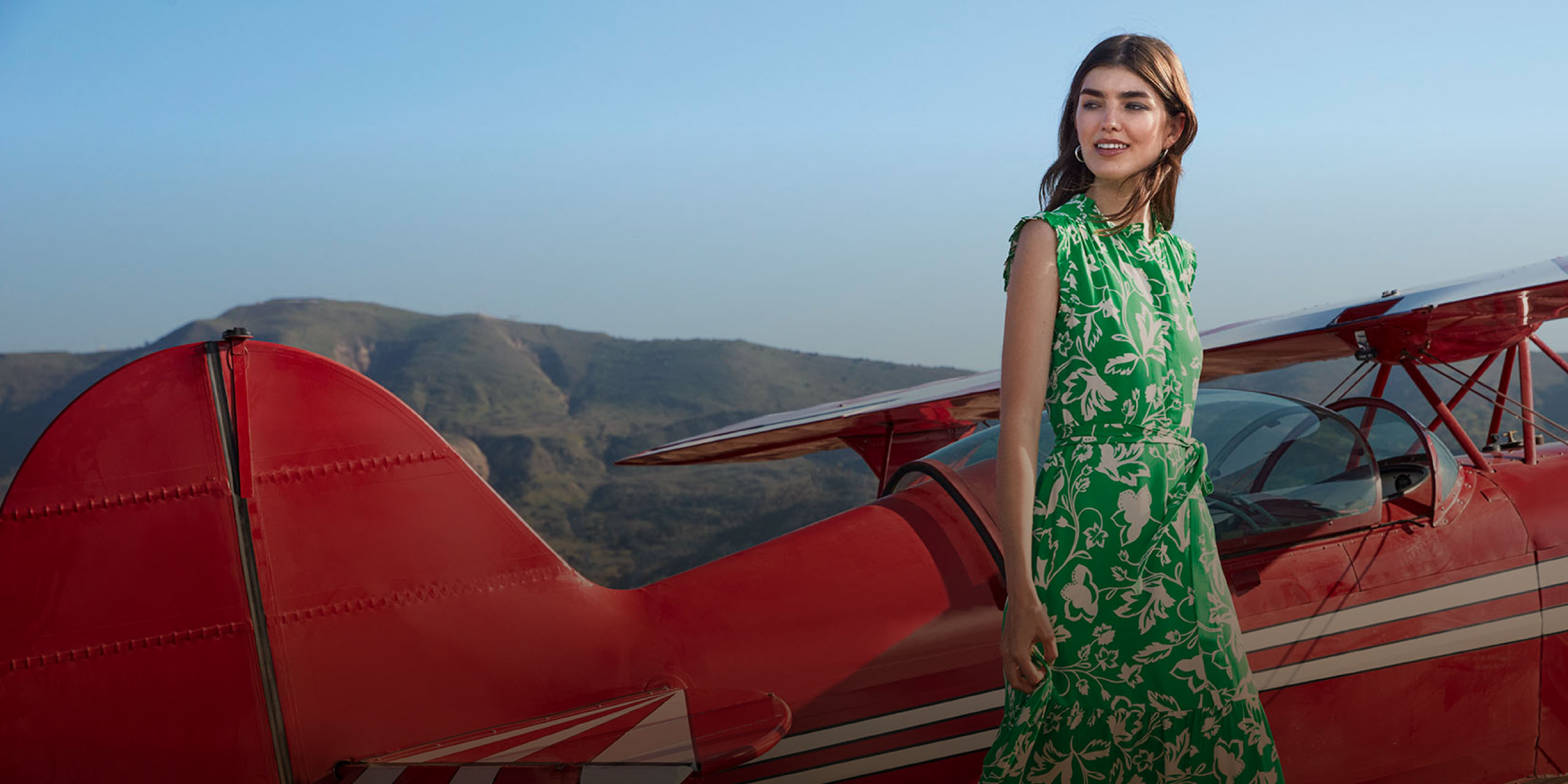 Image of model pictured in front of an aeroplane with a suitcase and wearing wedge sandals, a summer dress, sunglasses and a suede jacket.