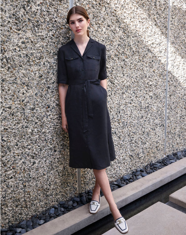 Image of model pictured standing by a wall wearing a tailored navy linen dress.