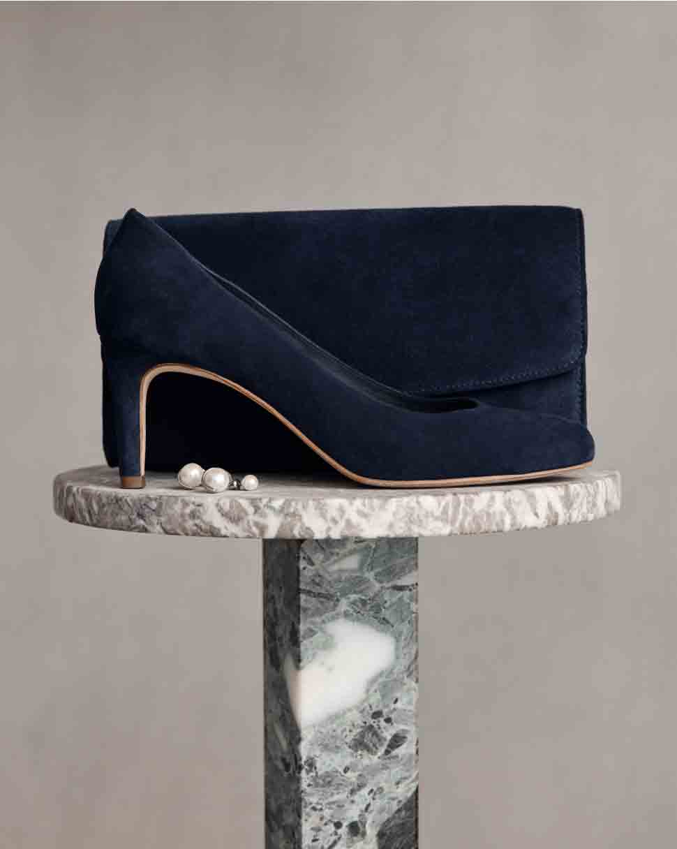 A navy suede court shoe and matching clutch bag sit atop a marble plinth