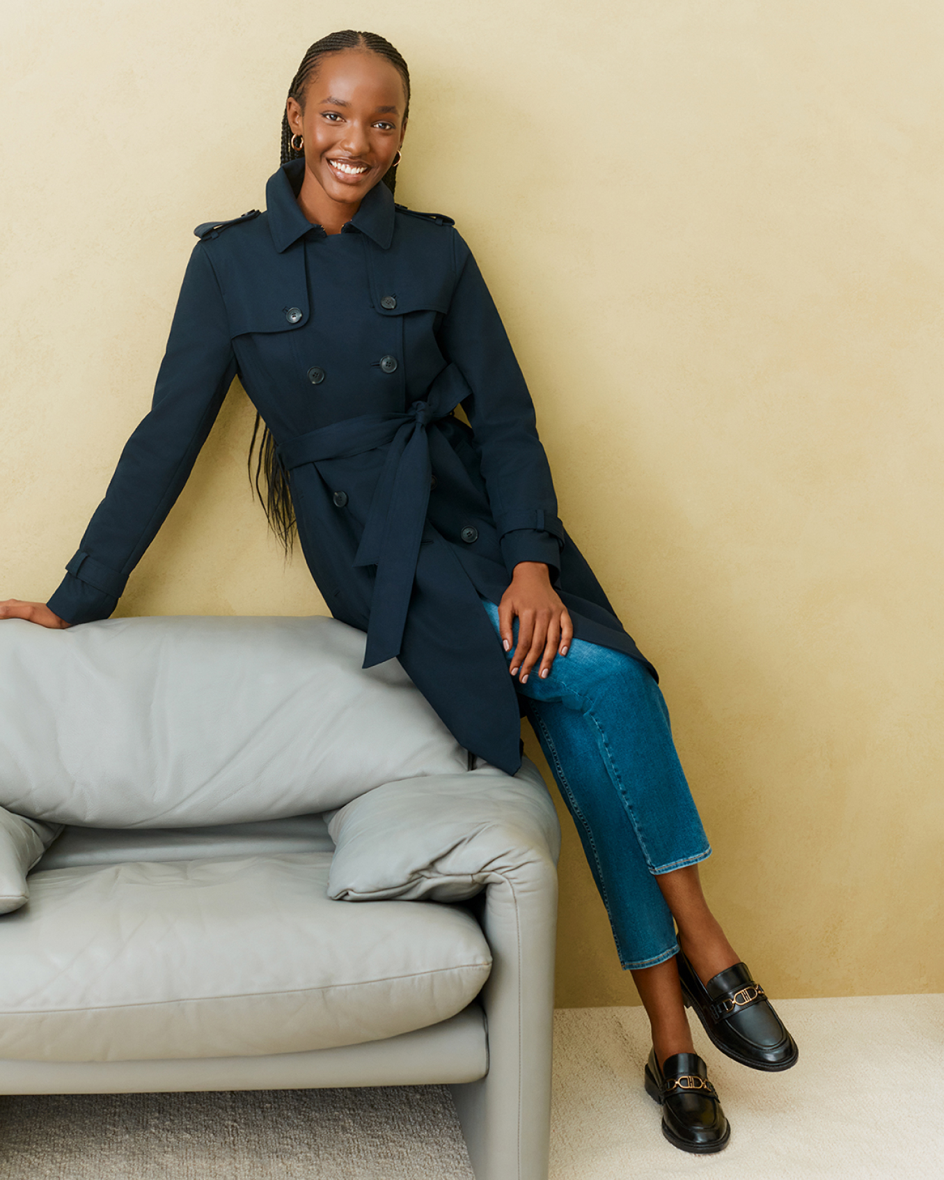 Hobbs models sit on the arm of a chair wearing a navy blue trench coat