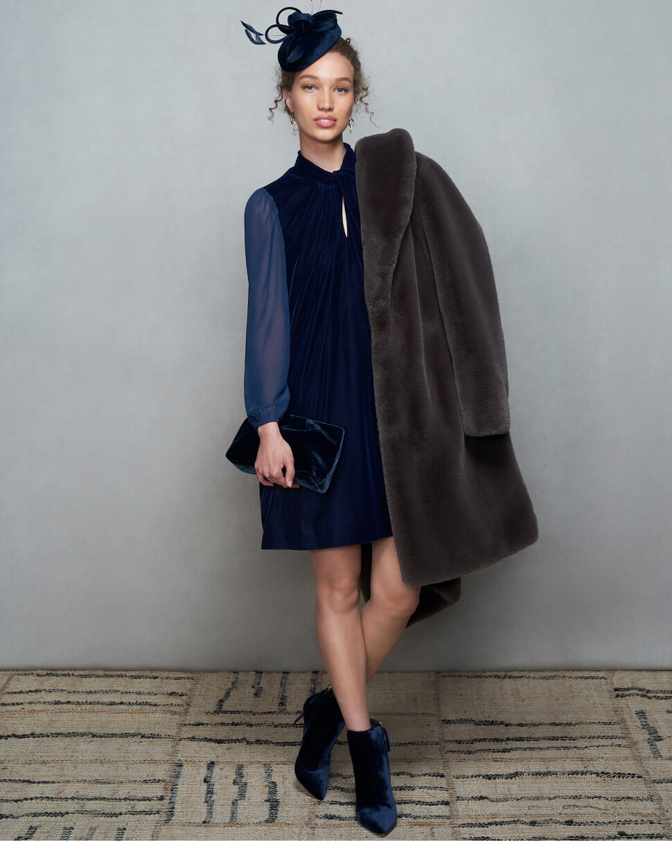 Model photographed against a canvas background wearing a Hobbs velvet tunic dress and faux fur coat draped over her shoulder.