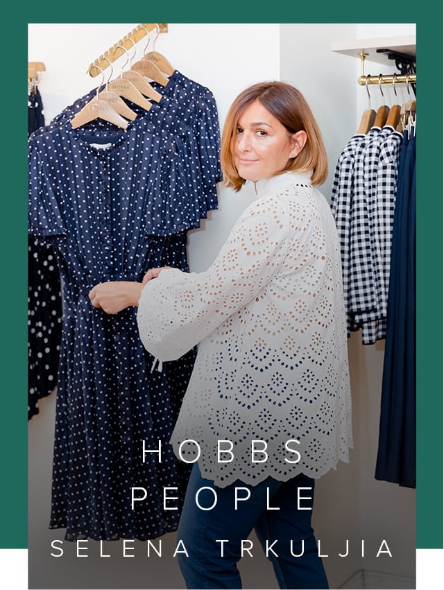 Hobbs UK & International Visual Specialist, Selena Trkuljia, photographed in a Hobbs store wearing a white blouse and blue denim jeans next to several blue dresses hung on a rail.