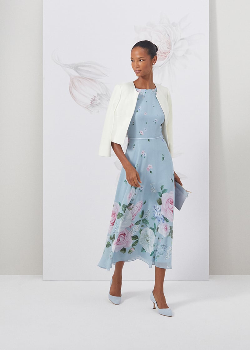 Image of model standing in front of a floral painted background wearing a floral dress with coordinating ivory blazer.