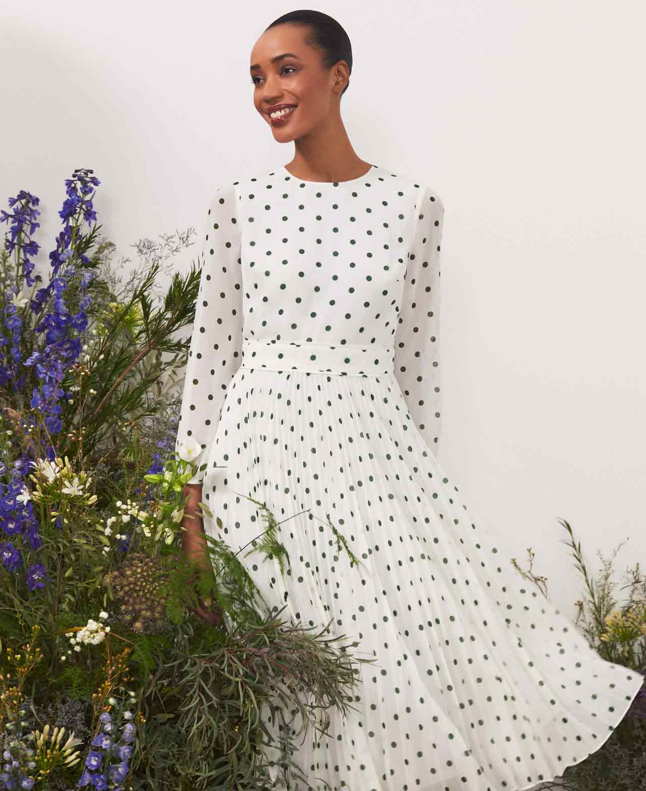 Hobbs stands next to a floral display wearing a polka dot fit and flare dress.