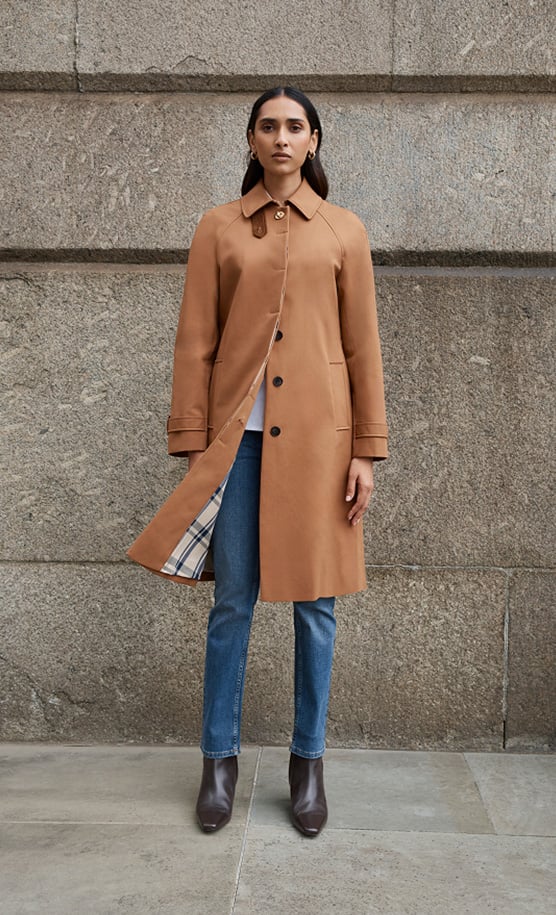 Model stands against a wall wearing a tan trench coat.