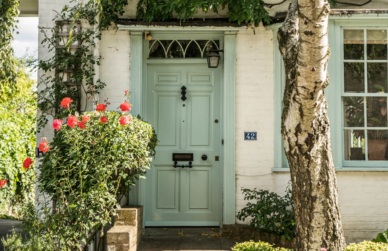 A serene pale grey green door with a picturesque pink flowers and green foliage surrounding it.