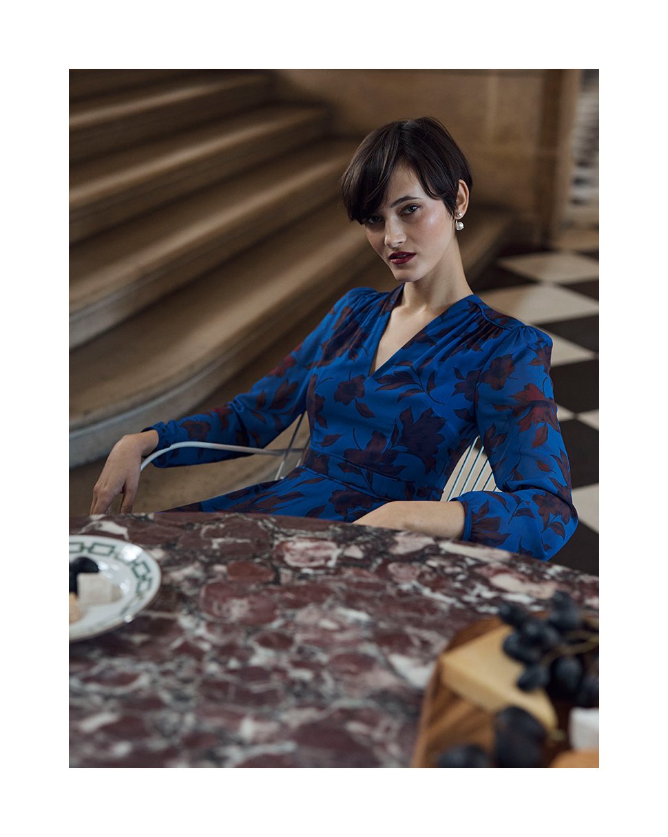 Hobbs model sits at a table in a blue floral print dress.
