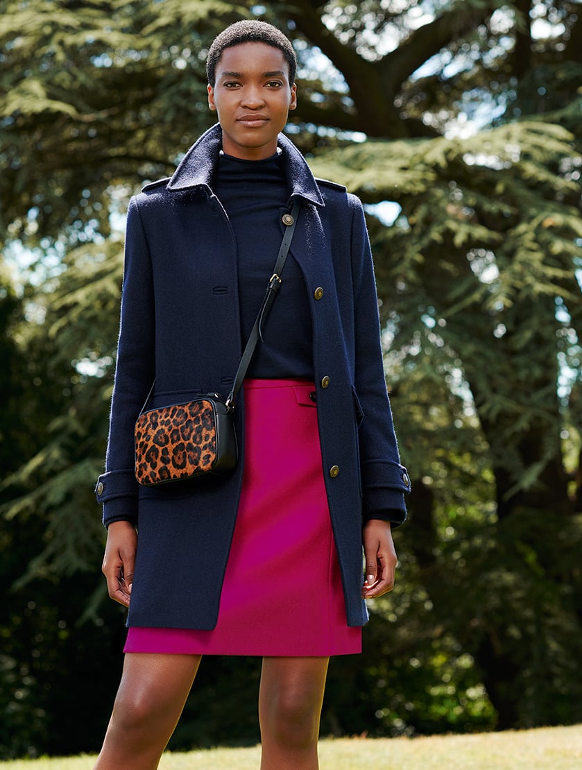 Alt text: Hobbs women’s winter wool coat in dark navy blue layered over a dark roll neck jumper and a pink skirt with a leopard print leather crossbody bag.