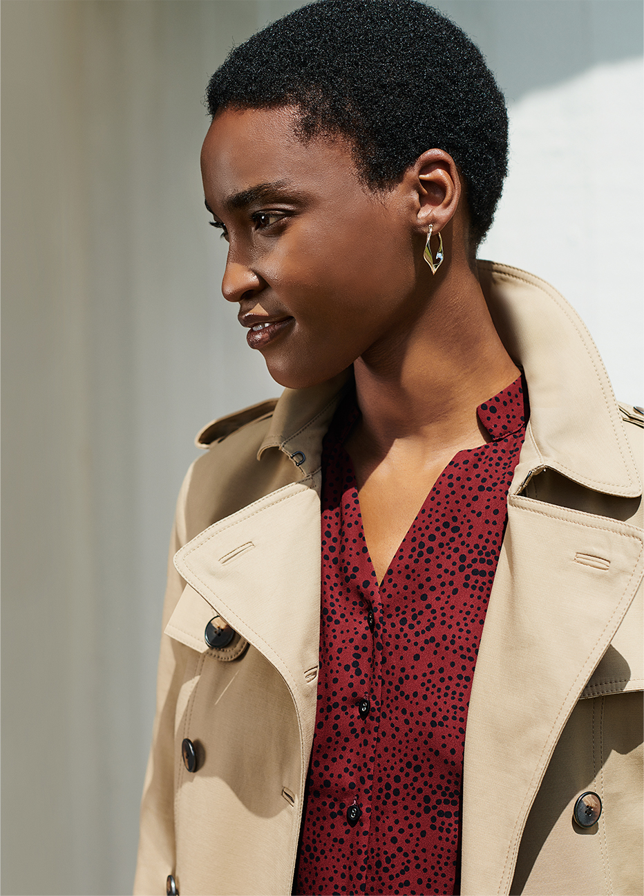 Hobbs model wearing a beige women's trench coat over a red shirt dress and hoop earrings.