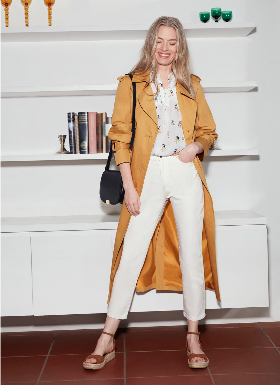 Model standing in front of a bookcase wearing a sandy yellow trench coat over a cream top and jeans.