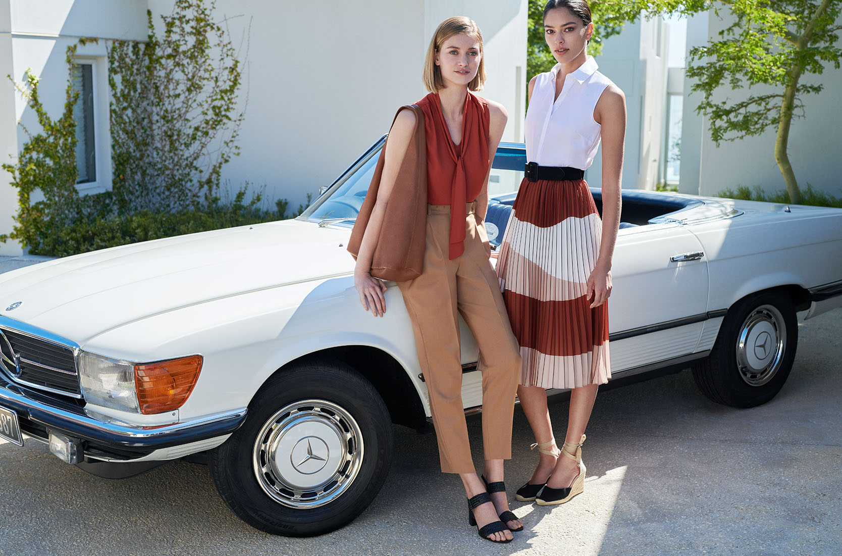 Two women pose in front of a white car, one wears a rust brown sleeveless blouse with tan tailored trousers with a black sandals and a brown tote bag, the other wears a white sleeveless blouse and a patterned brown, white, pink pleated midi skirt and black espadrilles.