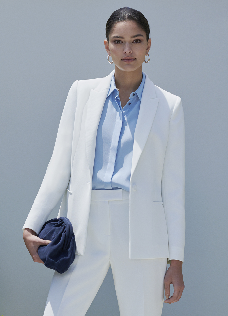 Women’s trouser suit in white worn with a pale blue collared shirt and a navy blue suede clutch by Hobbs.