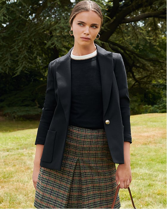 Hobbs contemporary women’s blazer layered over a dark jumper and check wool skirt, made with Abraham Moon’s British made sustainable wool fabric, worn with tights and styled with a brown leather tote bag