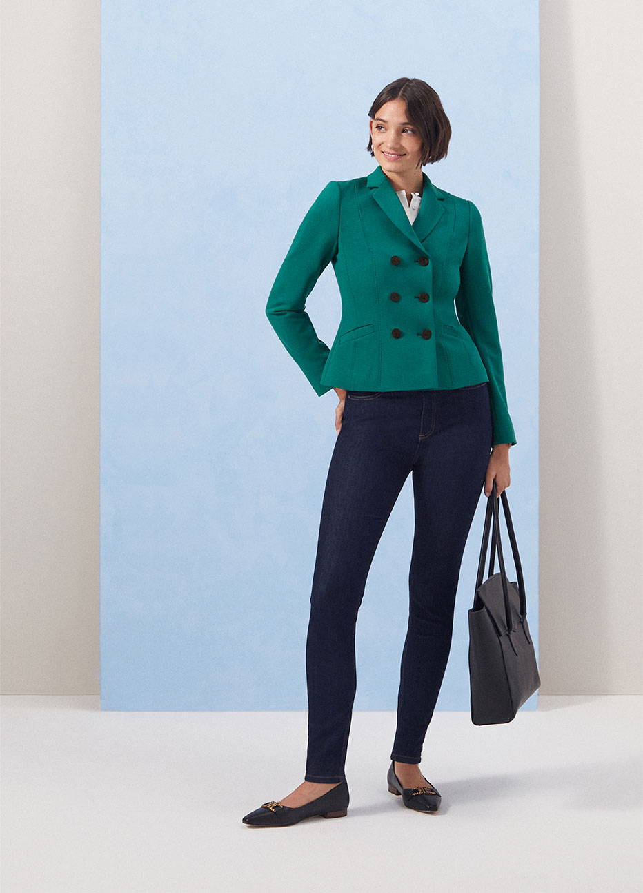 Model photographed against a blue backdrop wearing a Hobbs tailored jacket, white blouse and indigo jeans.