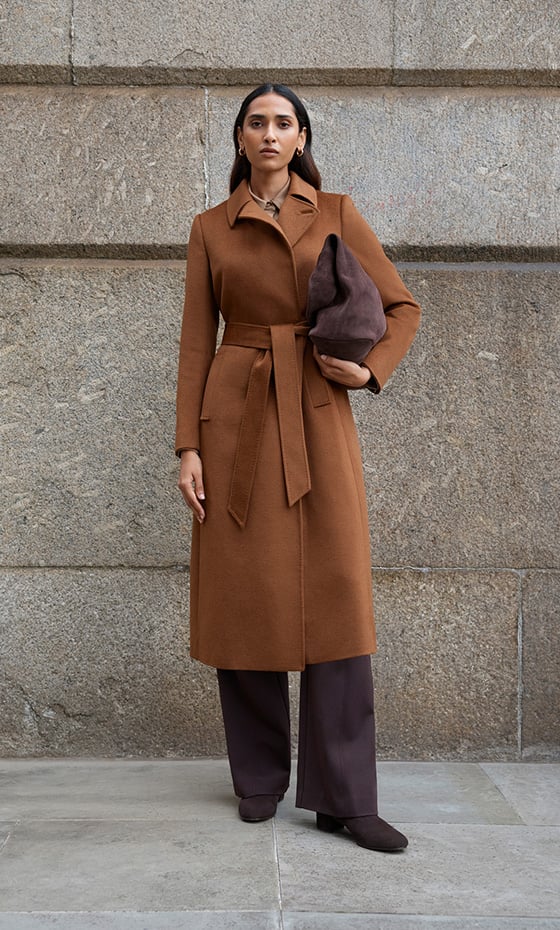 Model stands against a wall wearing a tan wool coat.