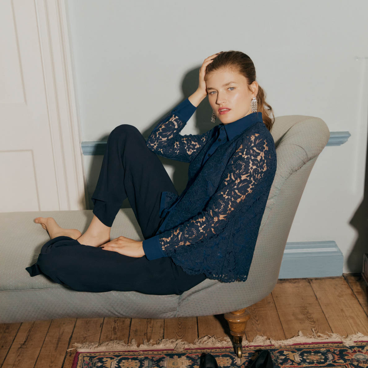 An image of a model sitting on a chaise longue wearing a lace top and smart trousers.