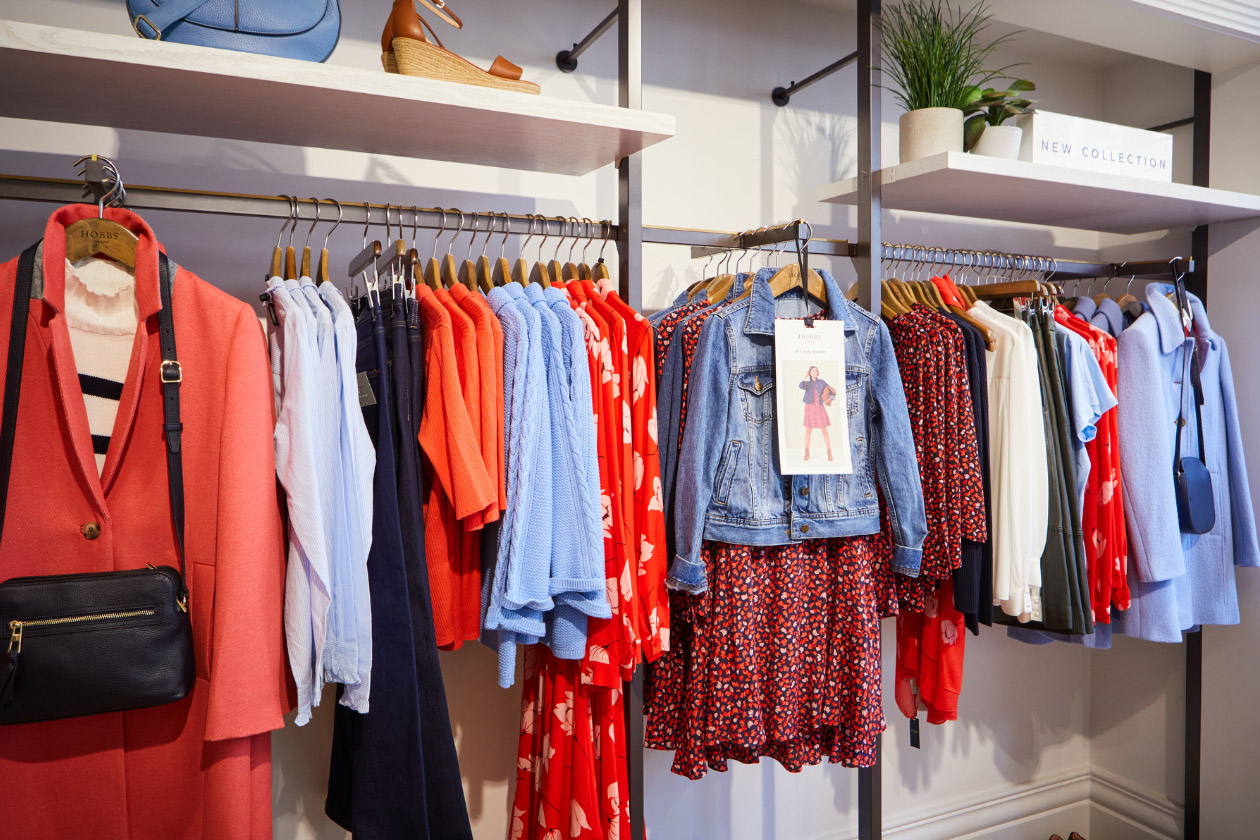 An image showing a section of a shelf in a Hobbs store. The shelf is decorated with a piece of framed artwork, a new collection sign, wrislets in blue, green, pink and white, court shoes in blush pink, and the upper half of a few hung up garments which include a dotted shirt in black and white, a polka dot top in black and white placed next to other designs in white, black and pink.