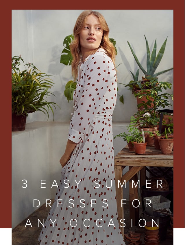 Model wearing a polka dot dress stands beside potted plants.