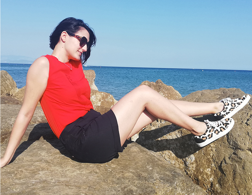 Marta Bonfaci, Senior Ecommerce Assistant at Hobbs poses on the rocks by the seaside wearing a red top, navy shorts, leopard print trainers and sunglasses.