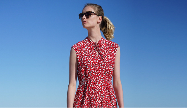 A Hobbs model wears a red dress with short cap sleeves and a neck fastening at the front, styled with sunglasses with blue sky in the background. 