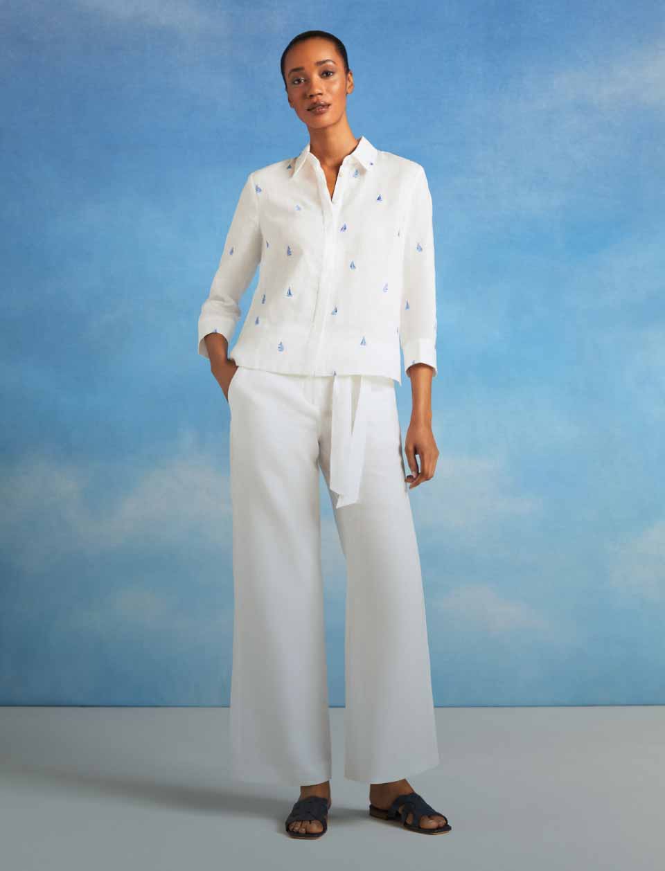 Model photographed in front of a sky blue background wearing a white linen shirt and trousers.