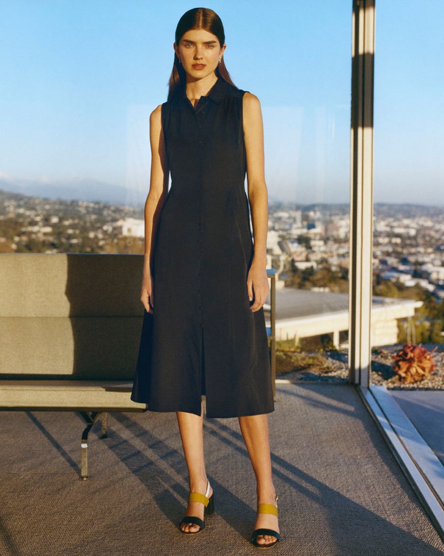 Image of a model pictured in front of a glass window and wearing a sleeveless summer dress and block heels.