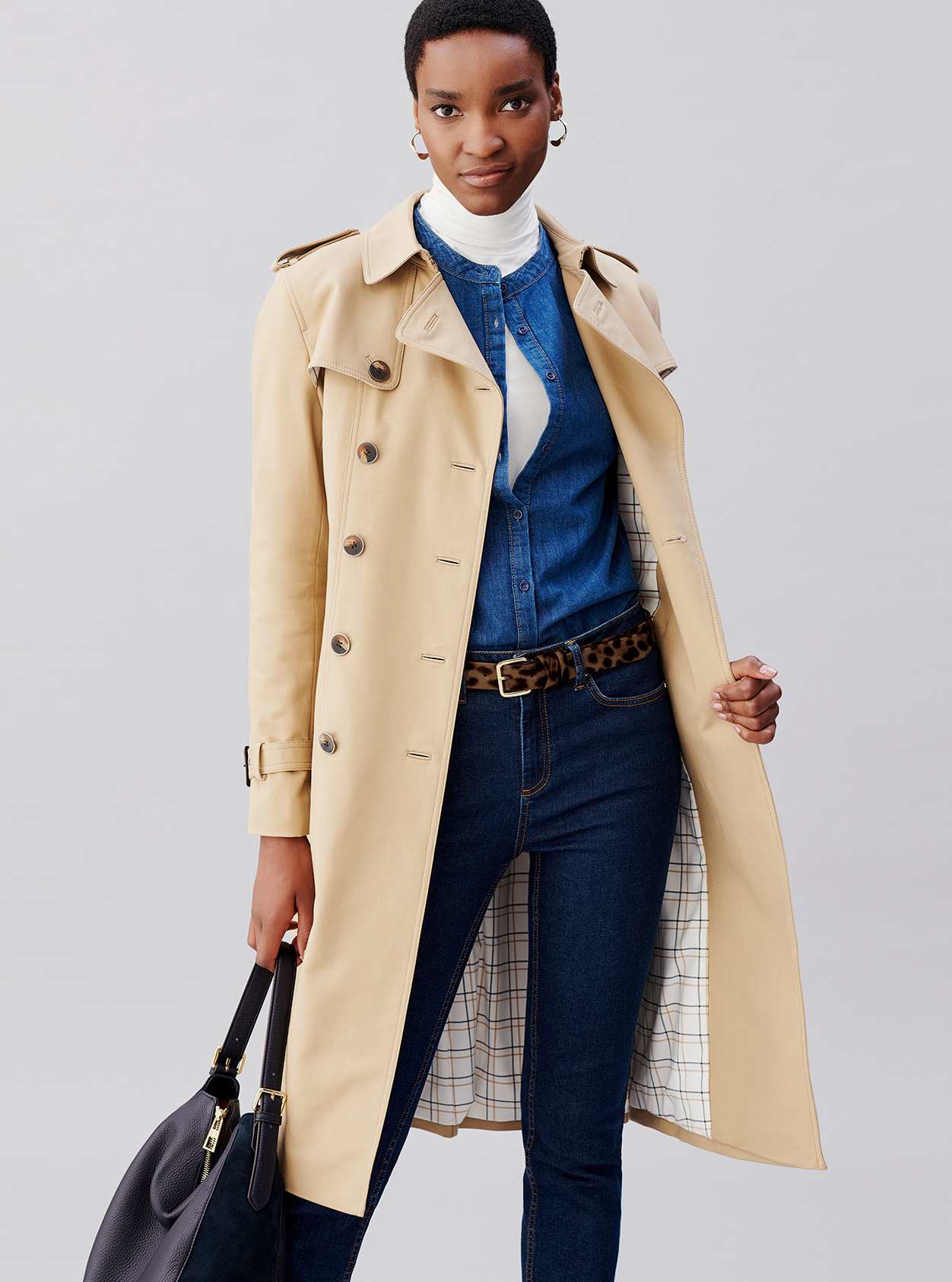 Hobbs beige trench coat for women layered over a denim shirt, white turtleneck top, blue denim jeans, leopard print belt and a black leather bag.