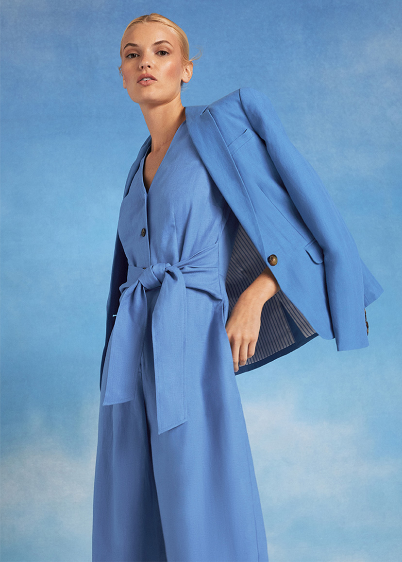 Close-up of model photographed against a sky background wearing a blue jumpsuit and white blazer.