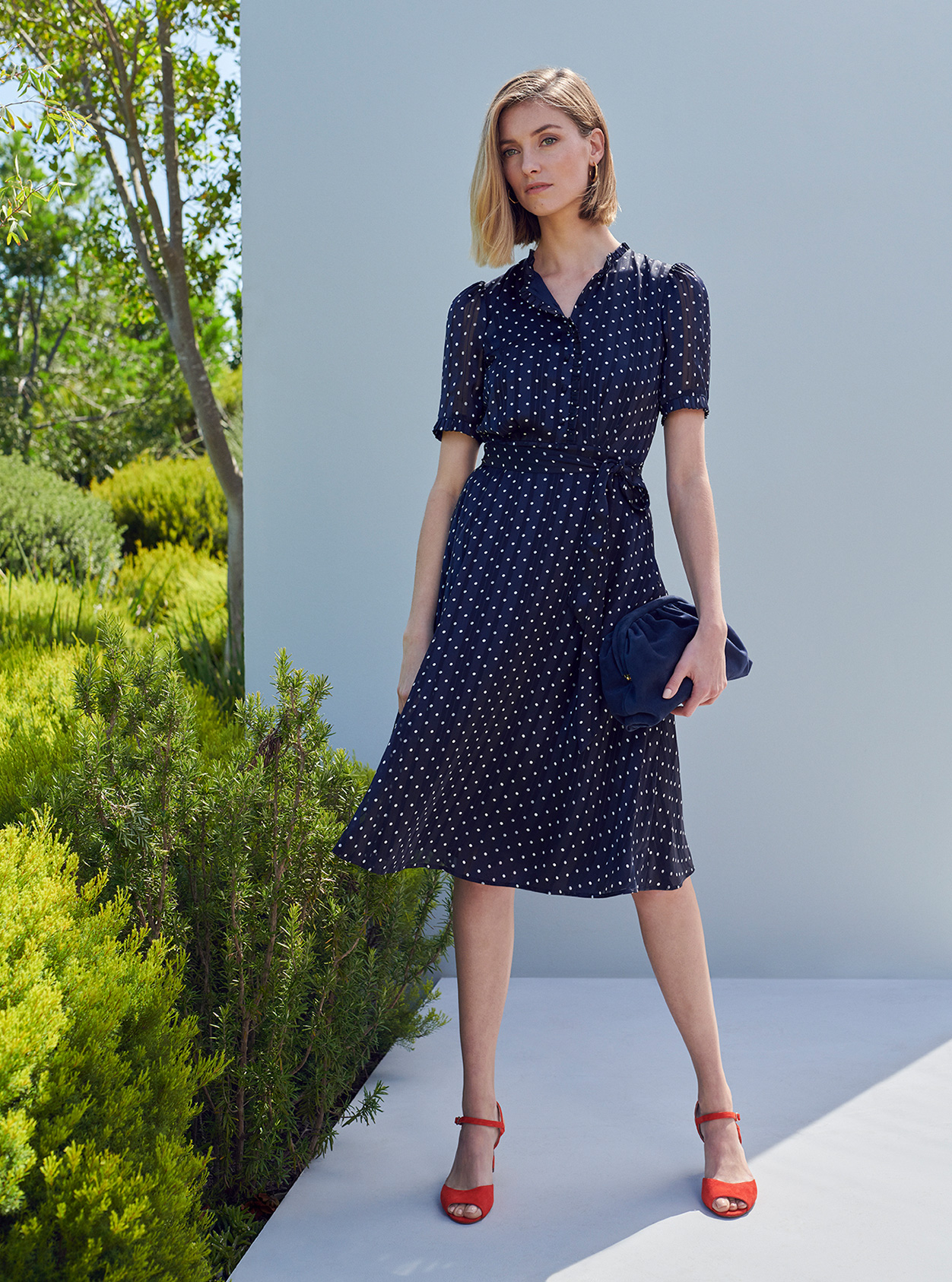 A woman poses in a navy blue and white spotty silk shirt dress with red heeled sandals.  