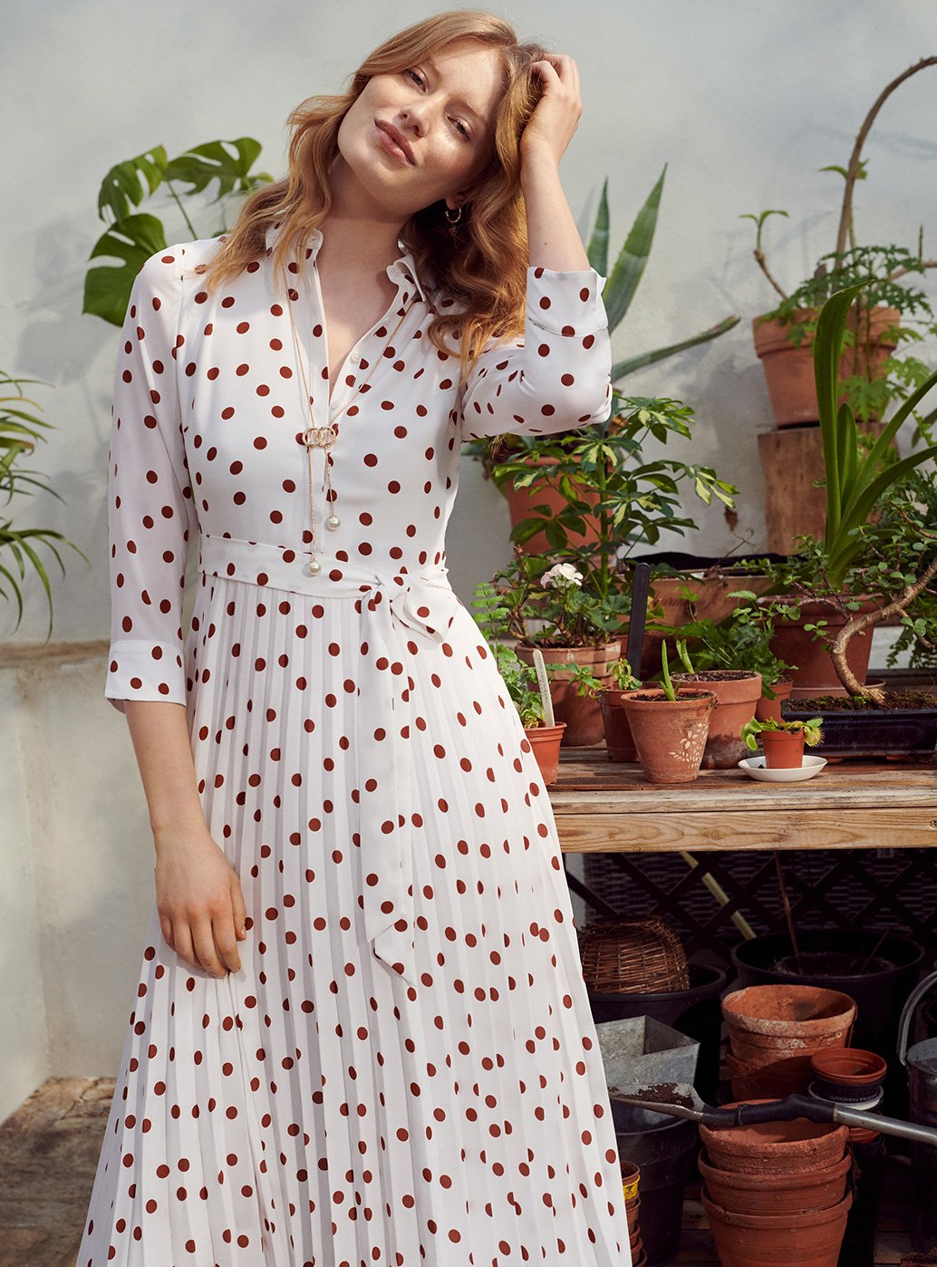 Model wearing a polka dot dress stands in front of potted plants.