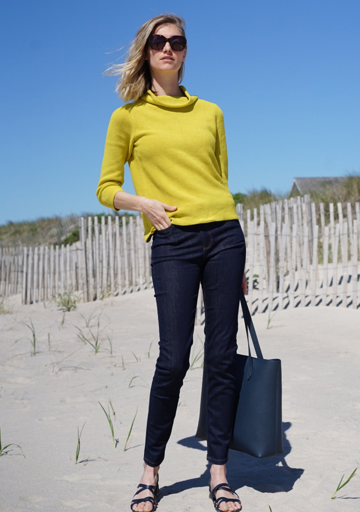 A blonde woman stands on a beach in a yellow roll neck jumper with dark denim jeans, sandals and a navy blue leather tote bags