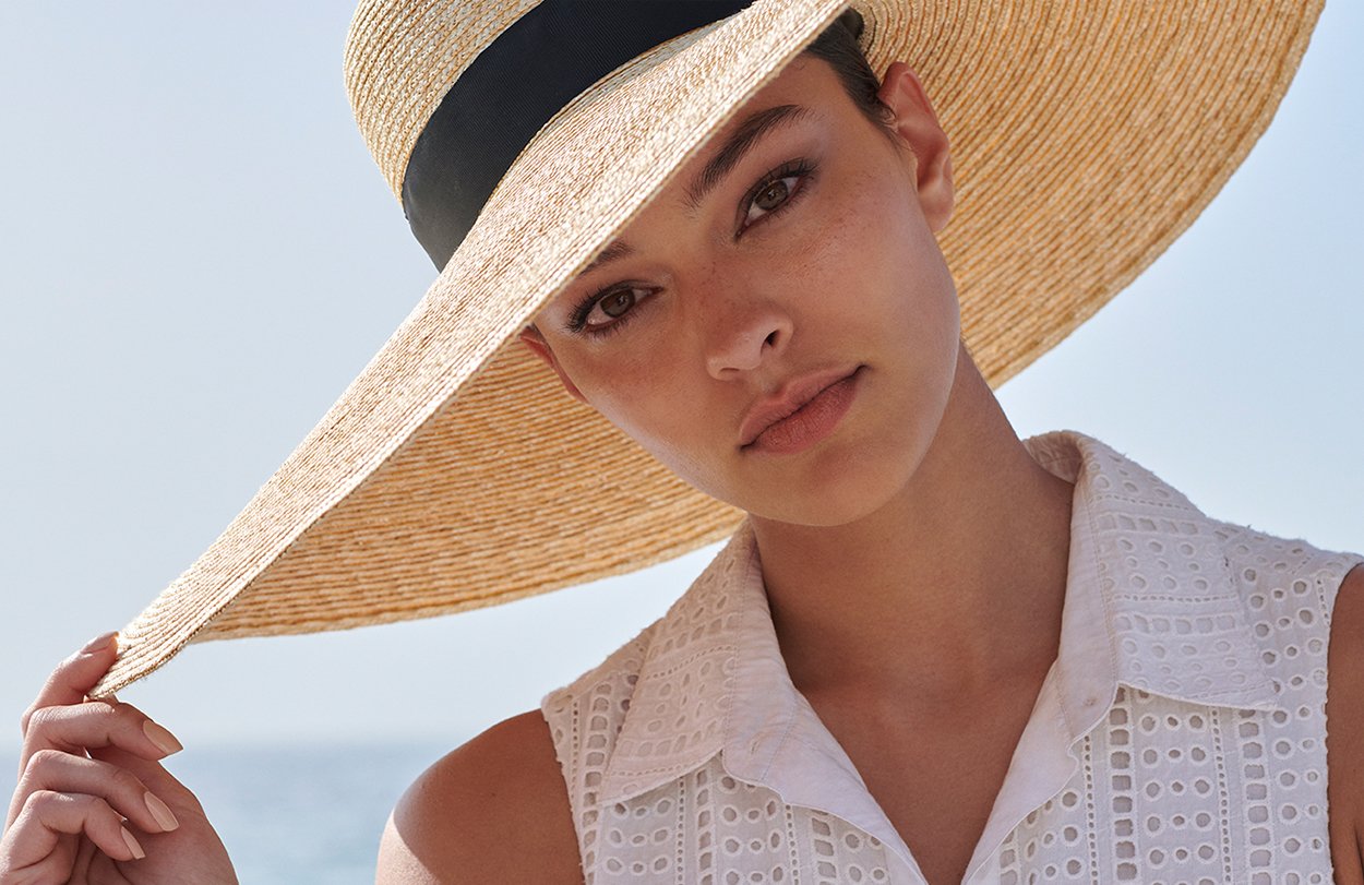 Dark haired woman wears a wide brimmed straw hat with a black band and wears a white broderie anglais sleeveless shirt