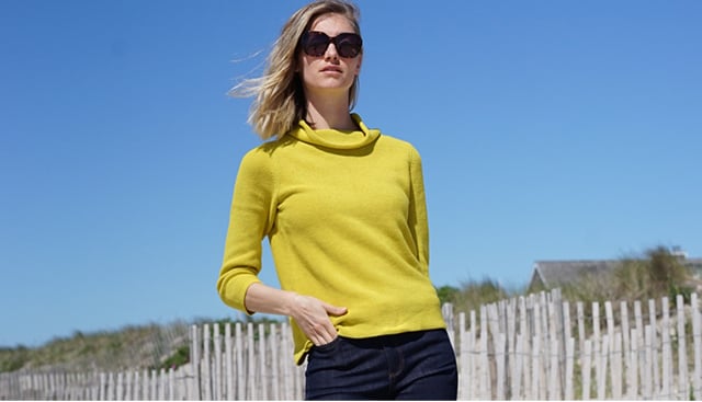 A woman poses in sunglasses, a bright yellow jumper wth dark denim jeans