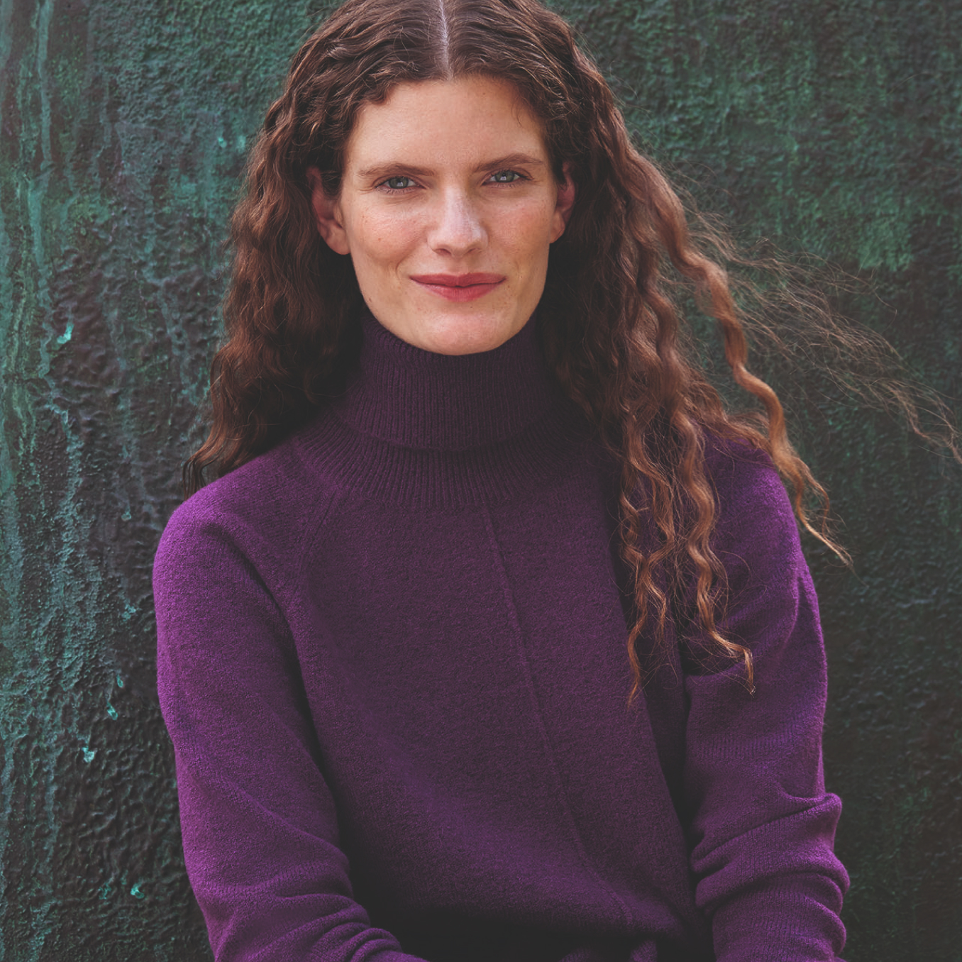 Alt text: Image of model sits against a patina sculpture wearing a purple cashmere jumper.