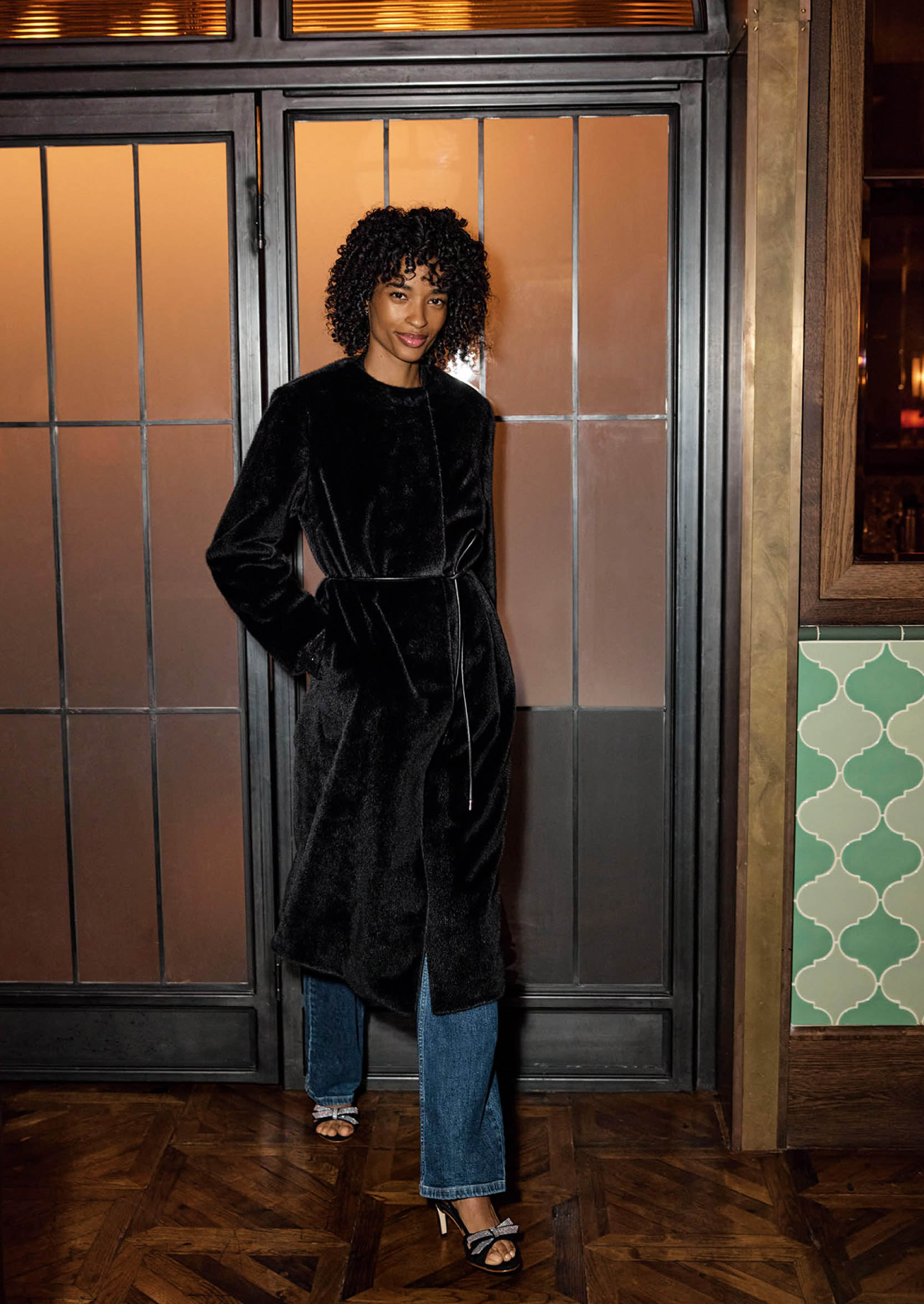 Model photographed standing up and wearing jeans, sandals, a shirt, a jacket and a coat.