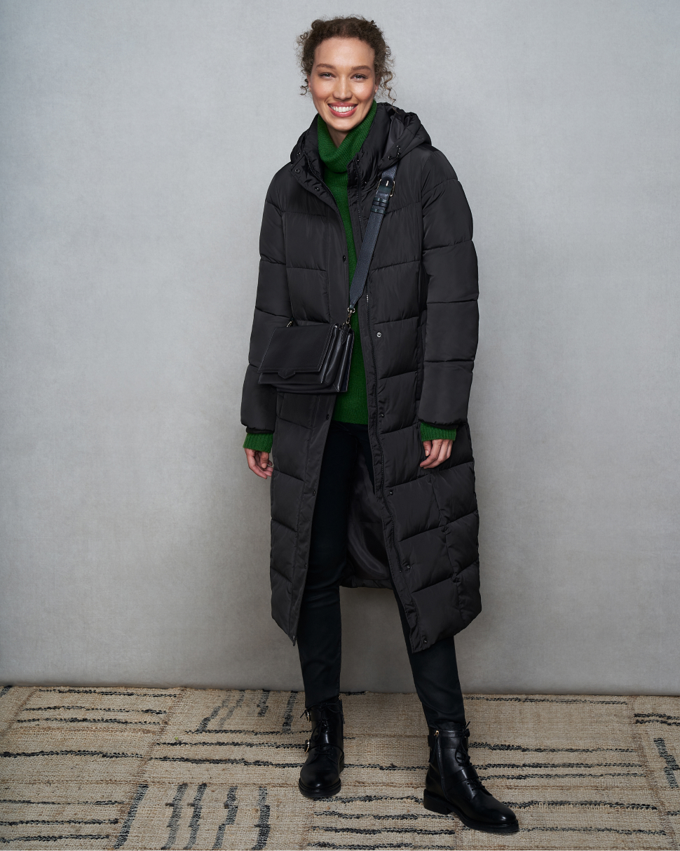 Model photographed against a canvas background wearing a Hobbs grey puffer jacket with green jumper, coated black jeans and chunky ankle boots.