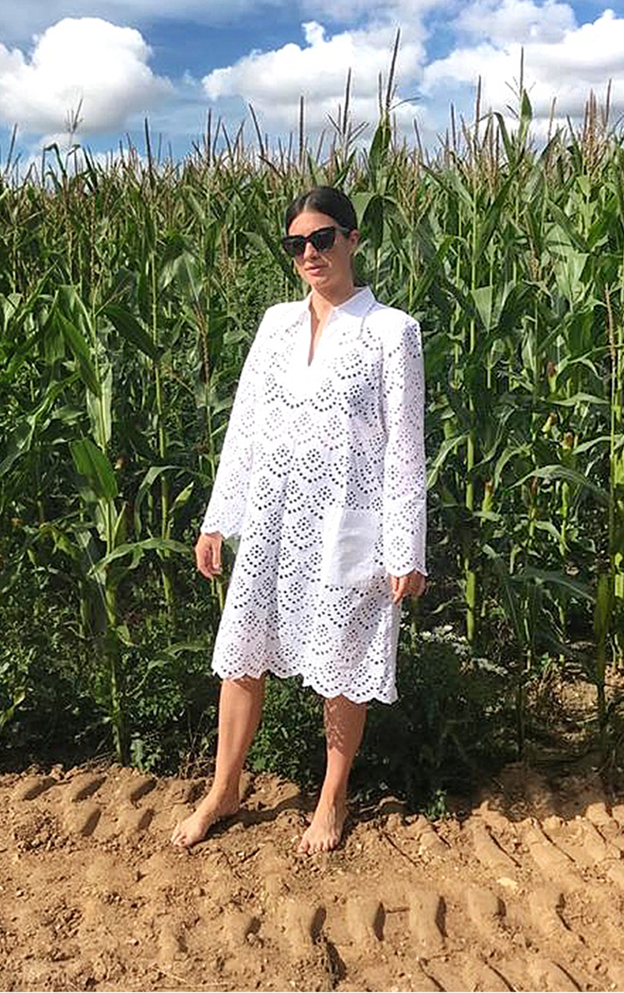Maddy Moxham, Brand Stylist at Hobbs stands in front of a wheat field barefoot wearing a white shirt dress and sunglasses.