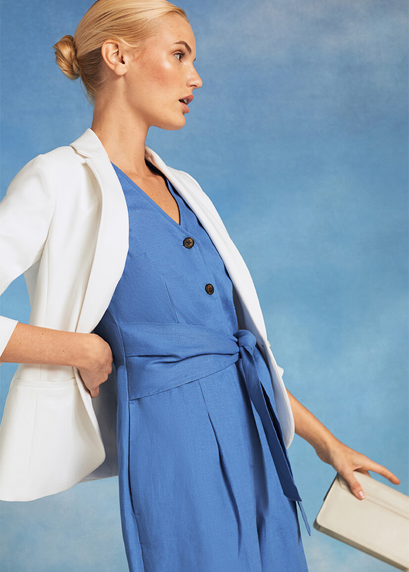 Close-up of model photographed against a sky background wearing a blue jumpsuit and white blazer.