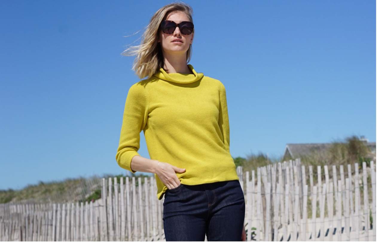 A woman poses in sunglasses, a bright yellow jumper wth dark denim jeans