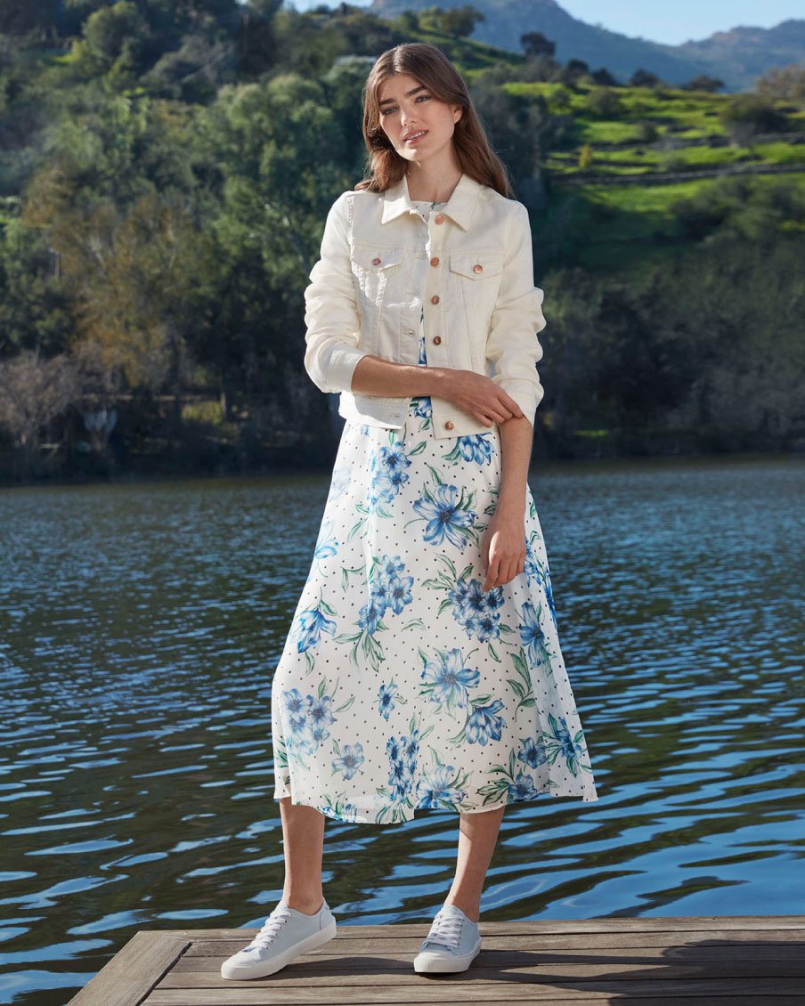 Image of a model pictured in front of a lake view wearing a floral summer dress,a  denim jacket and trainers.