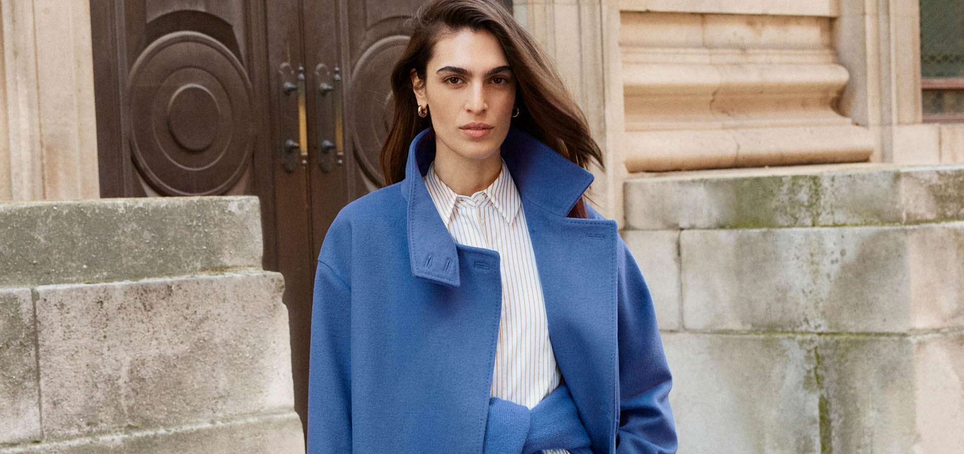 A woman wearing a blue jumper and coat, layered over brown trousers.
