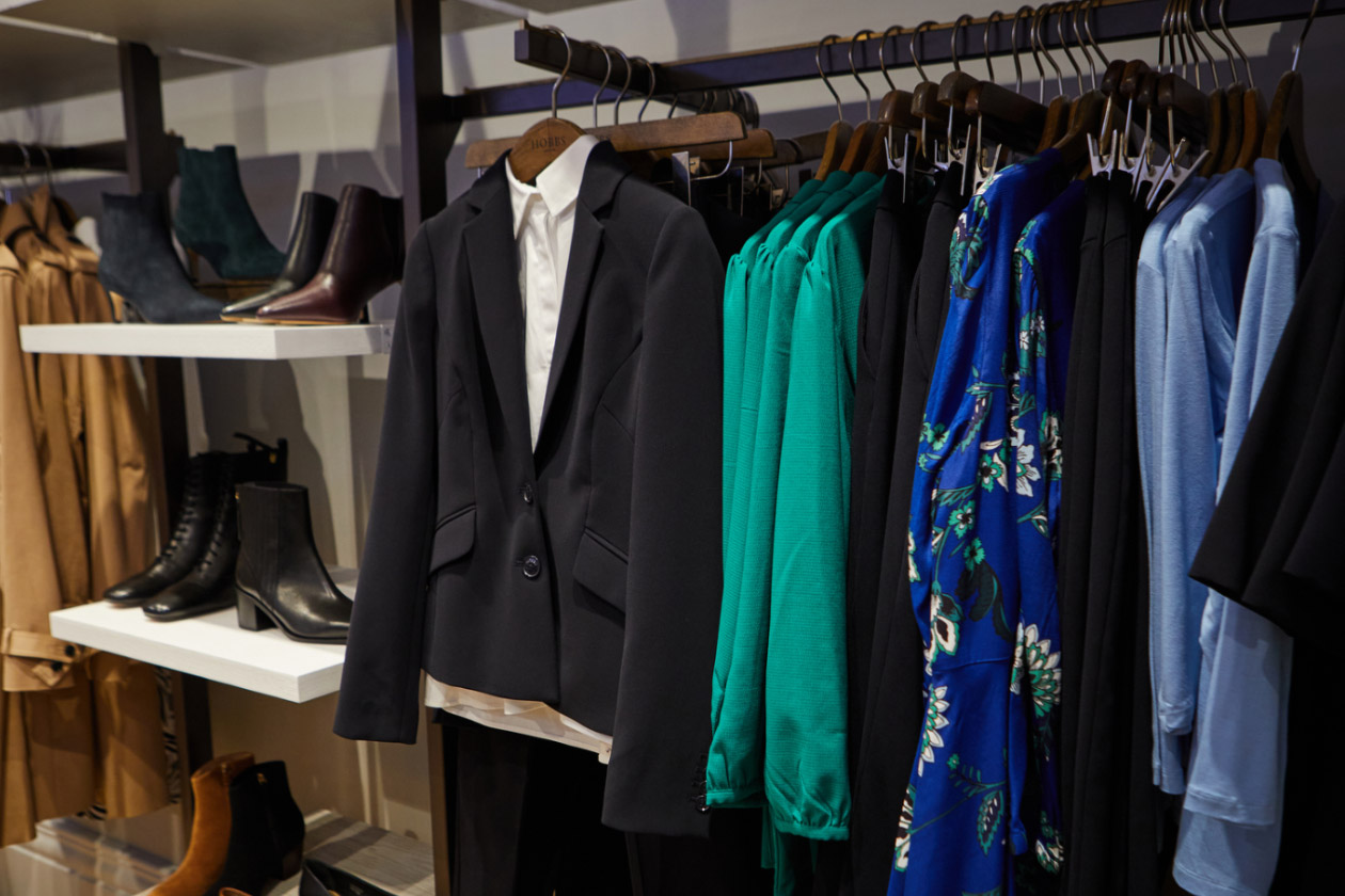 A clothing rail in a Hobbs store. The hanger in the middle shows a dogtooth patterned shirt dress in black and white with a crossbody leather bag in white. The hangers on either sides feature garments in black, white, grey and red.