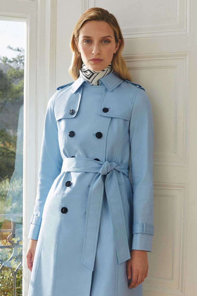 Dark haired woman poses in a green trench coat, white shorts & navy trousers holding a navy blue leather handbag.