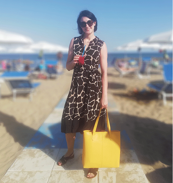 Marta Bonfaci, Senior Ecommerce Assistant at Hobbs poses on the rocks by the seaside wearing a red top, navy shorts, leopard print trainers and sunglasses.