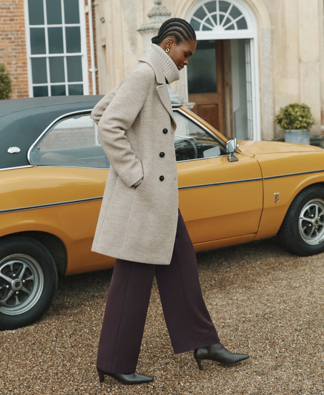 A woman wearing a beige coat layered over a high neck jumper and brown trousers.