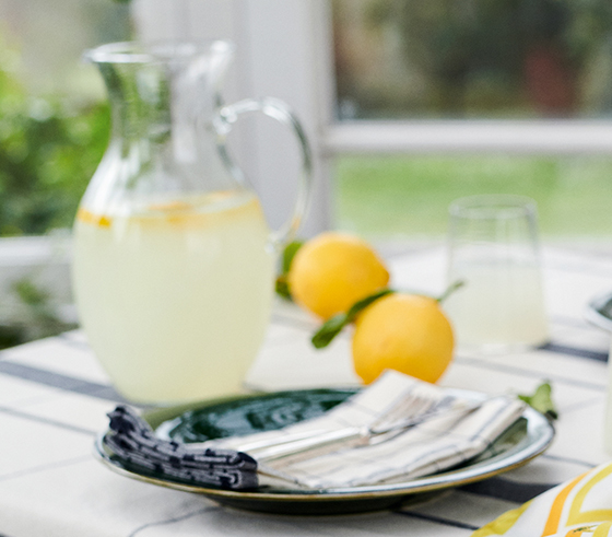 A jug of homemade lemonade, bowl of fresh lemons and a silk scarf sit atop a table.