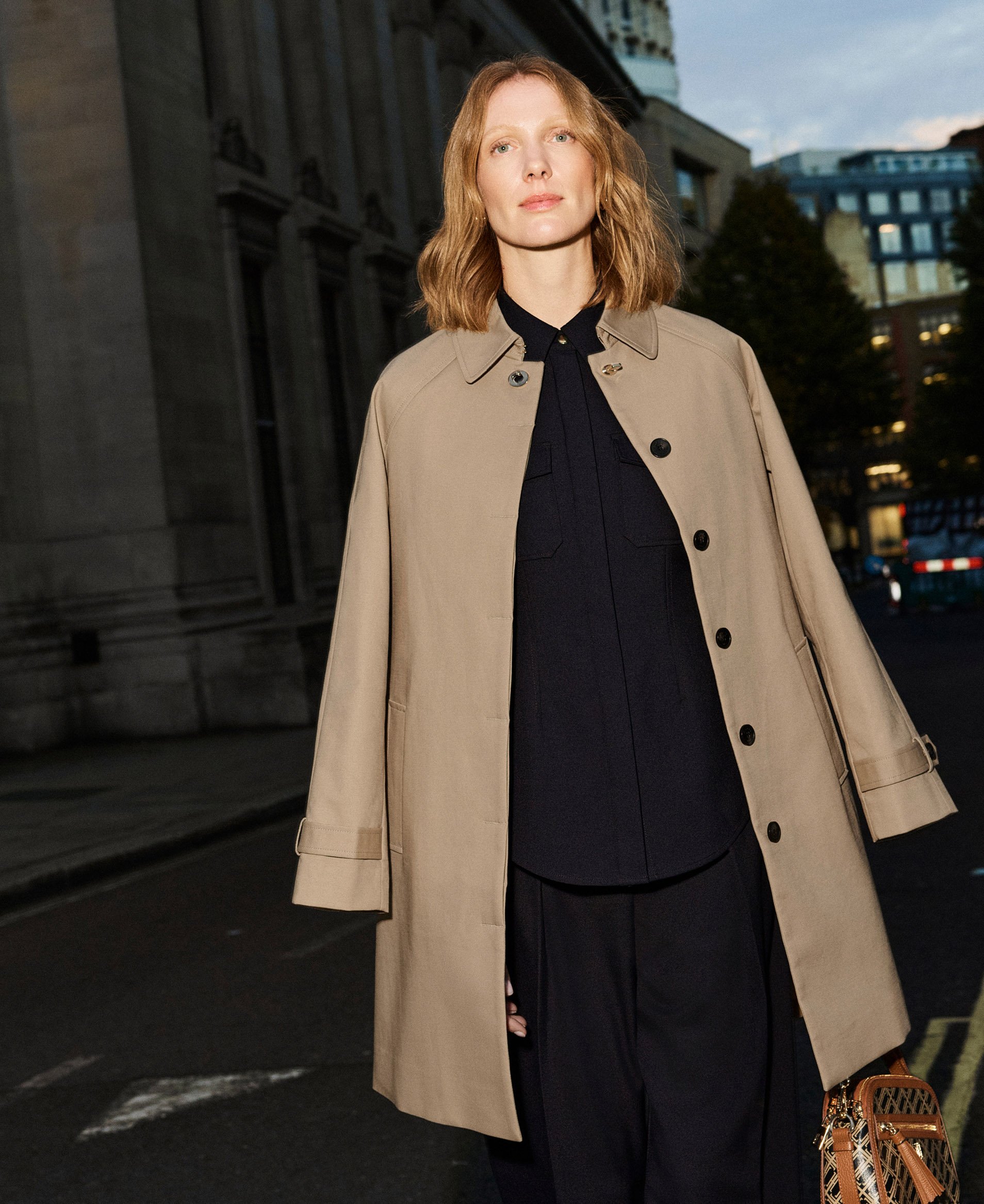 A woman wearing a camel trench coat into the evening.