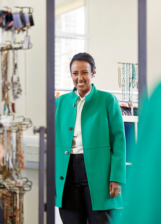 Woman Smiles At Her Reflection Wearing A Bright Green Coat, White Shirt And Dark Tailored Trousers.