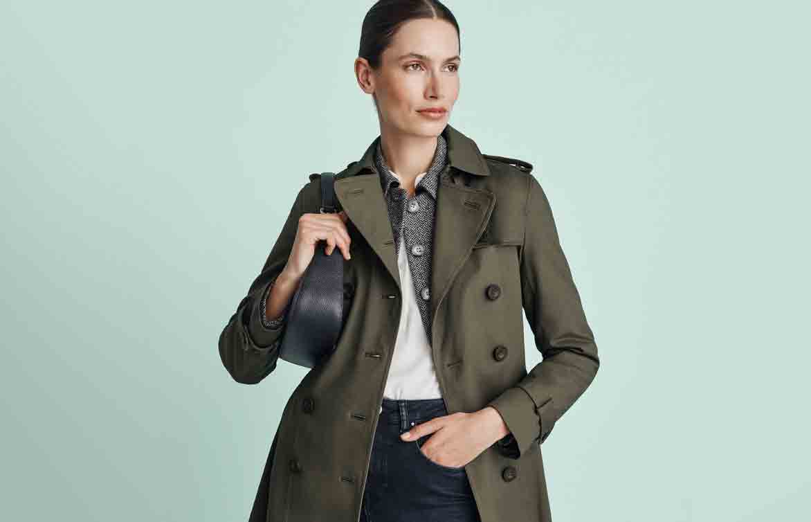 Hobbs model wears green trench coat and jeans.