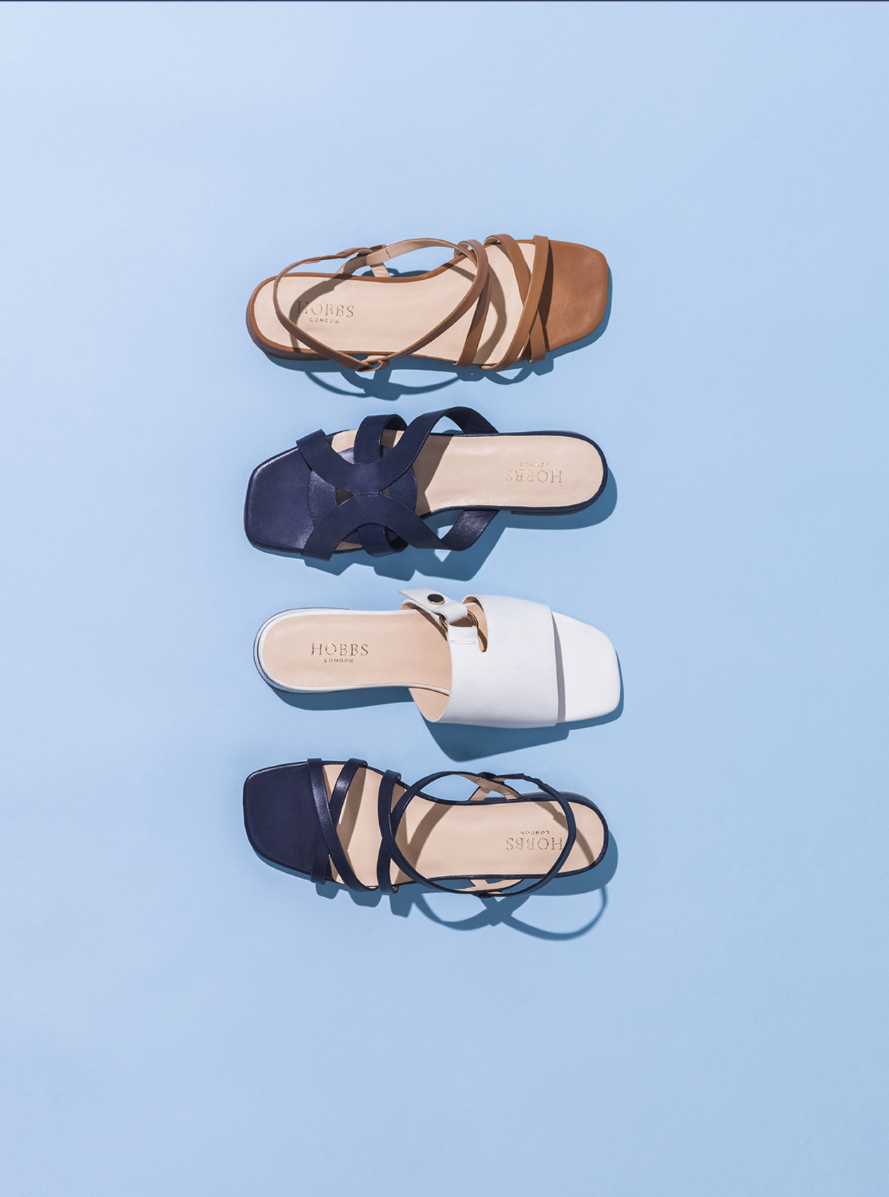 Four summer sandals from Hobbs against a blue background, from top to bottom: Brown sandal, dark blue sandal, white sandal, black sandal.